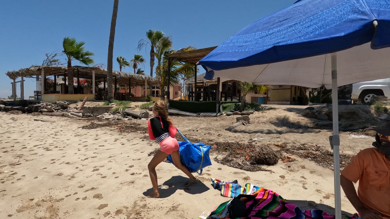 Child turns with a large blue bag beside a blue umbrella on the beach.
