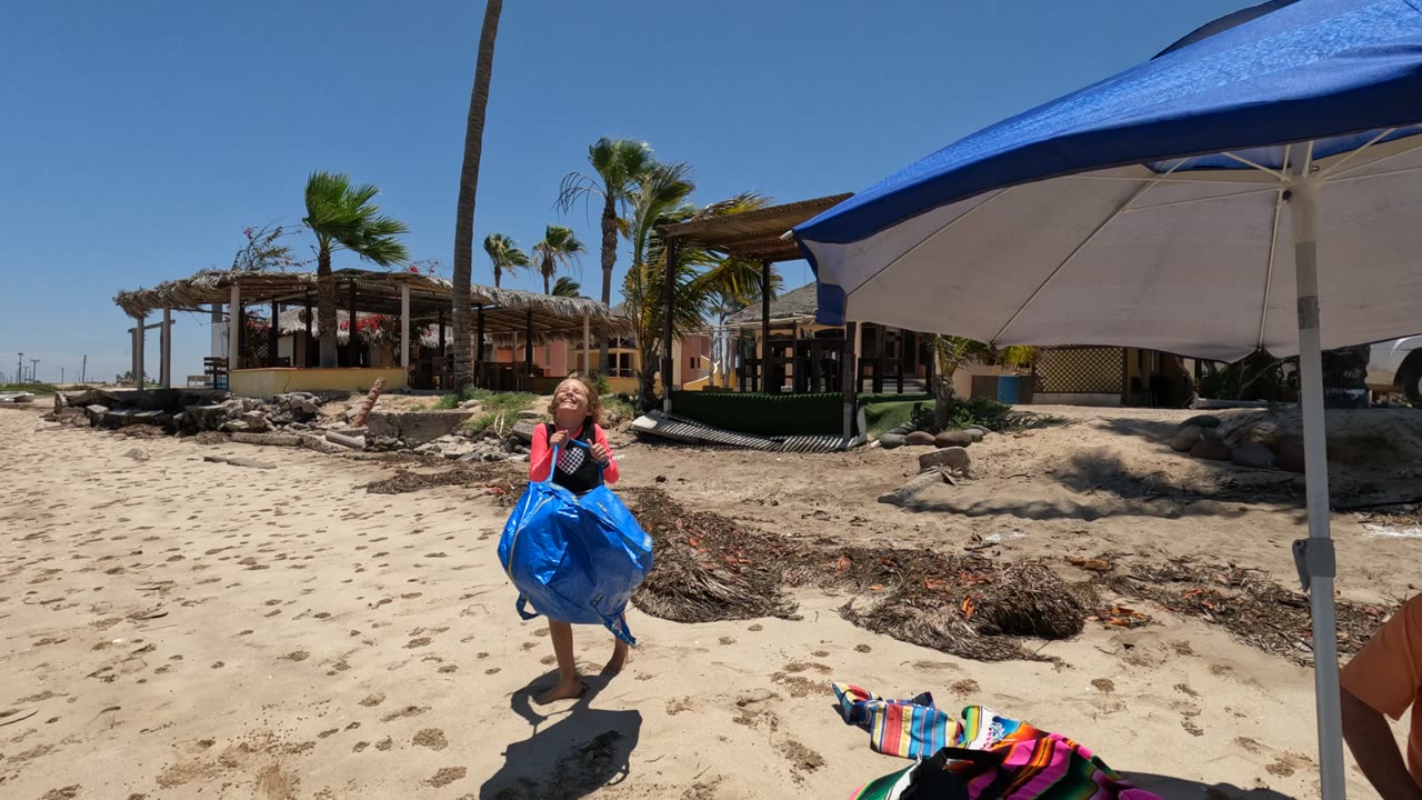 In Baja California Mexico, the child carries the blue bag farther from the umbrella across open sand, with the shoreline, palms, low beach buildings, rocks, and a colorful towel visible around the frame.