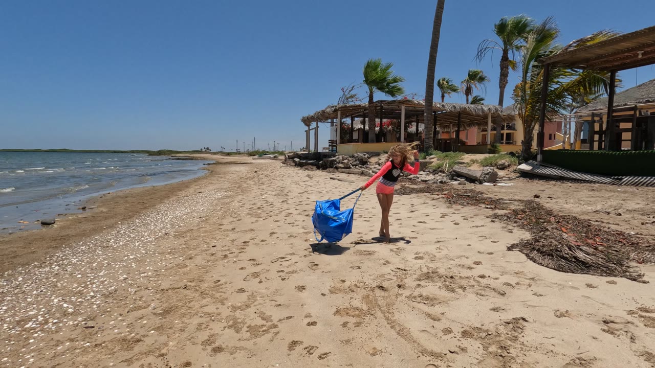 Girl in a pink top bends near a large blue bag on a sandy Baja California Mexico beach, with ocean water on the left and palm trees and thatched structures behind her.