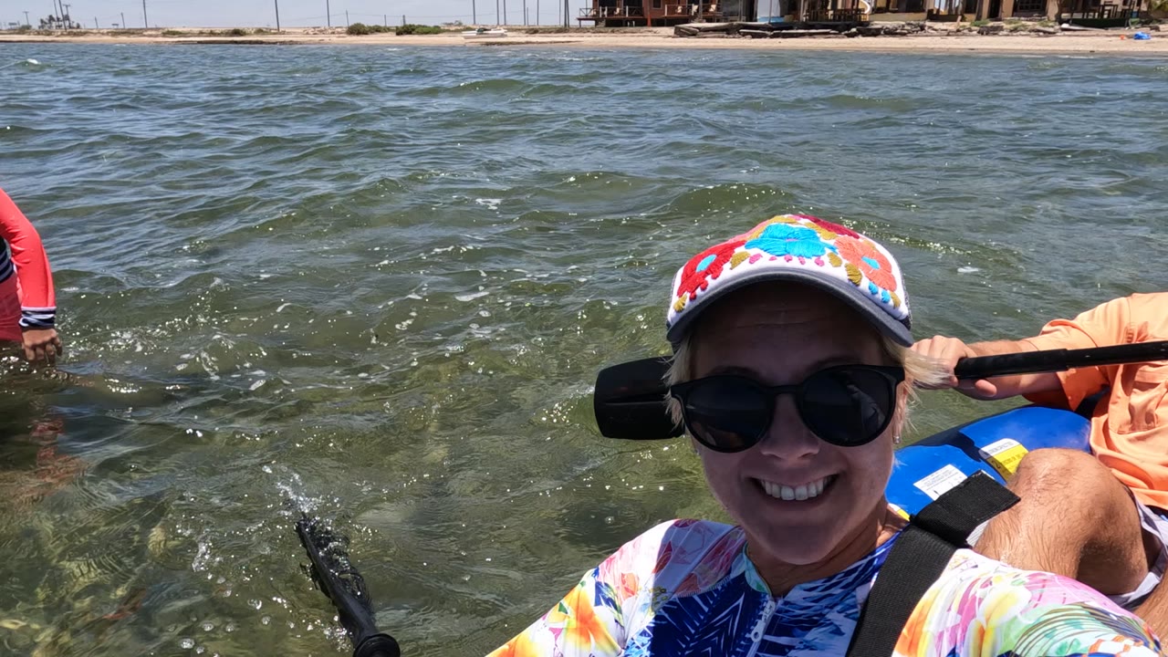 In Baja California Mexico, a smiling woman in sunglasses fills the foreground of a blue inflatable boat while a man behind her holds a paddle on green water near a sandy shoreline.