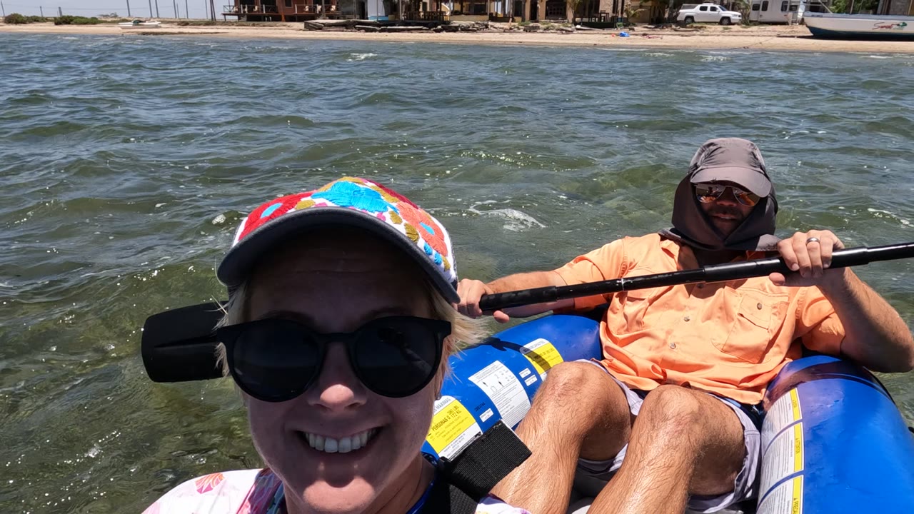 A tighter Baja California Mexico boat selfie shows the woman's face and cap at left foreground, the man seated behind with a paddle across the frame, and beach houses along the distant shore.