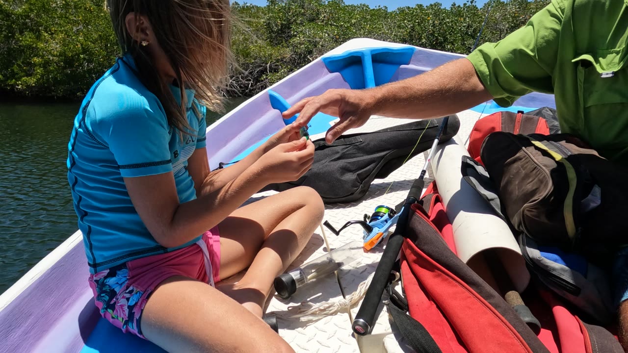 In Baja California Mexico, an adult in a green shirt reaches in from the side as the girl looks down at the small fish on the boat, with fishing gear and shoreline trees in the background.