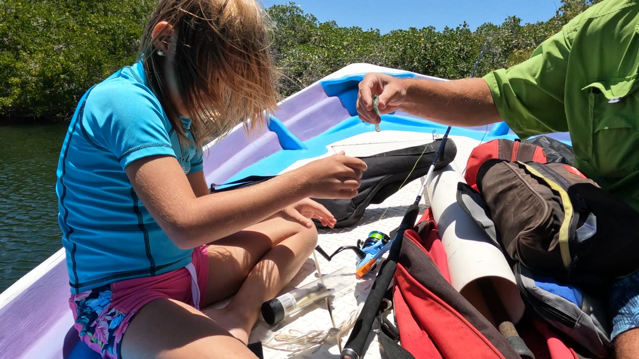 In Baja California Mexico, the girl leans over the small fish while an adult hand points toward it, with a red bag, dark backpack, fishing rod, and calm water behind them.