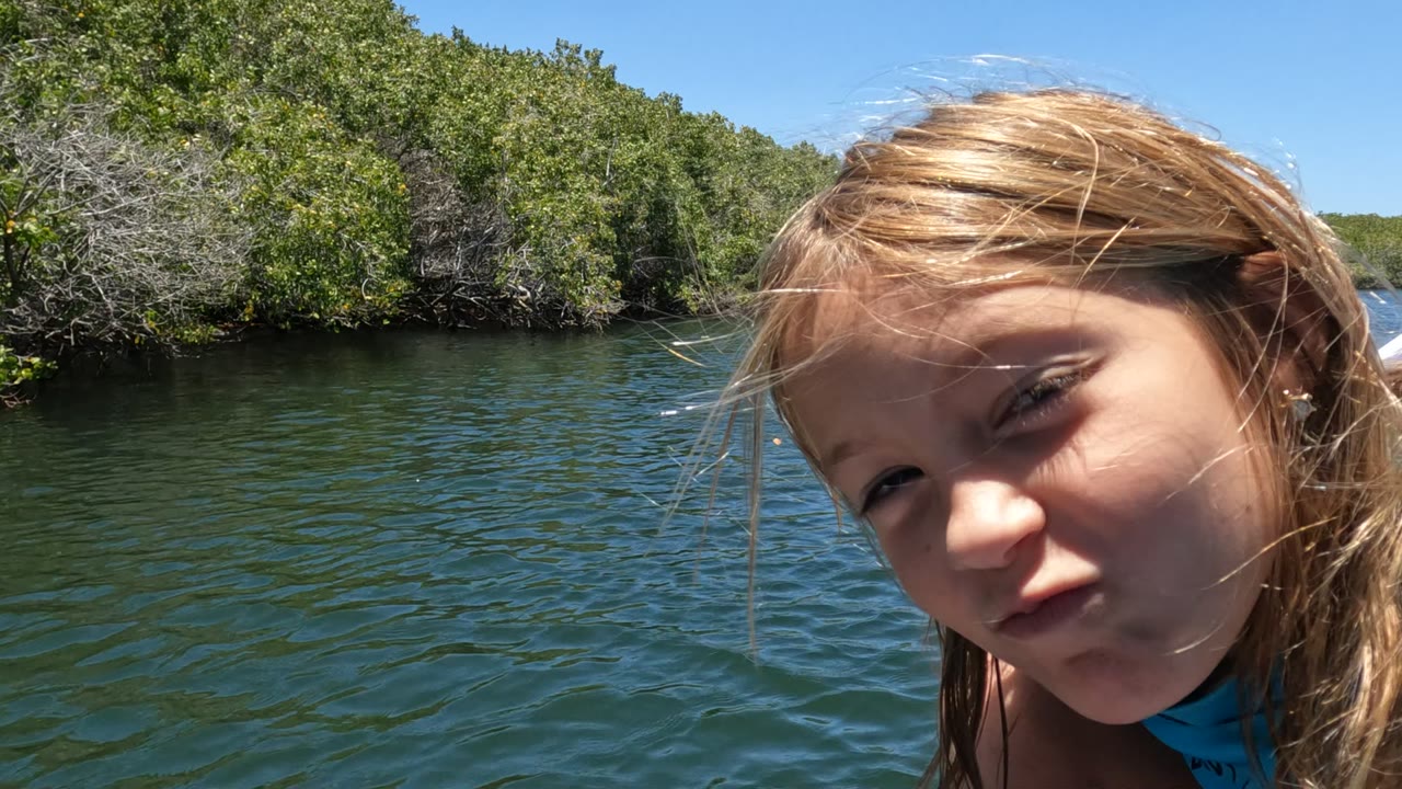 The girl's face fills the right side of the Baja California Mexico frame while green water and a continuous mangrove shoreline remain visible behind her in bright sun.