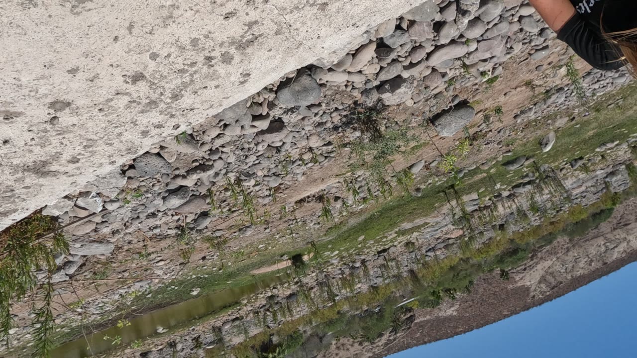 Opening frame of a girl partly cropped at the edge of a Baja California, Mexico canal scene, with peeling wall, rocks, sparse greenery, water, hillside, and blue sky visible.