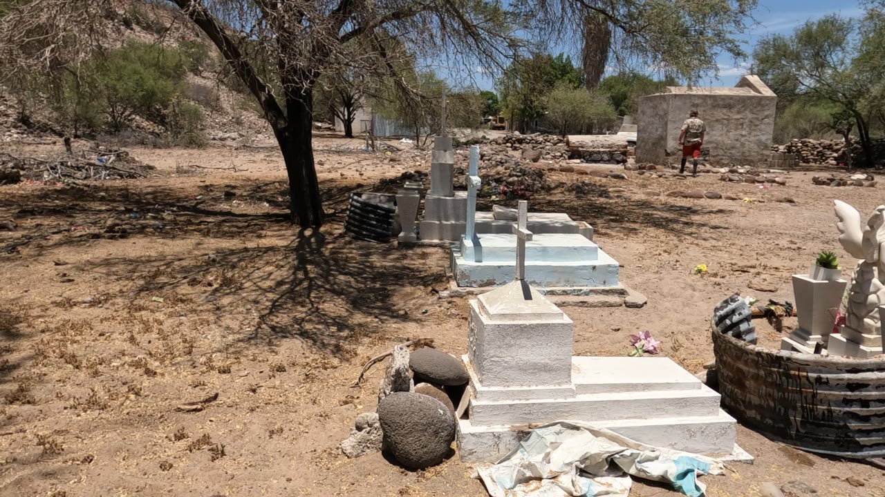Baja California, Mexico cemetery with white tombs and crosses in the foreground, dry sandy ground, sparse trees, and a distant person near a wall.