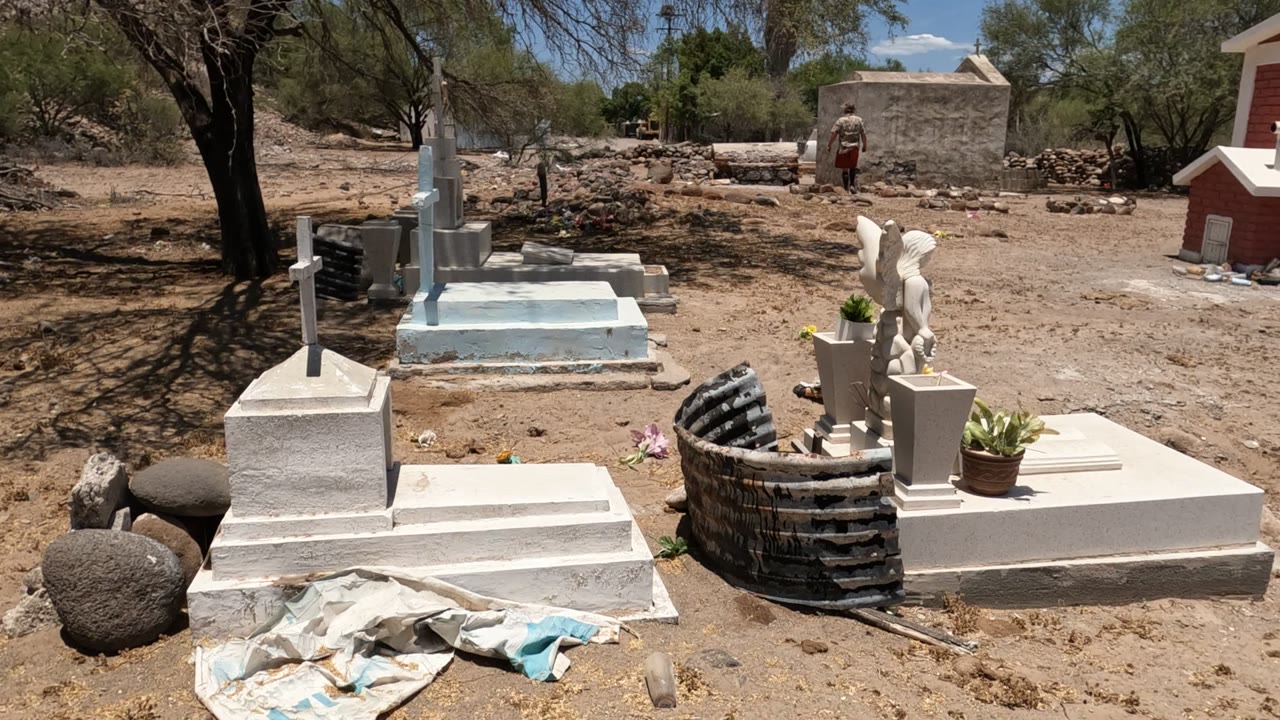 Baja California, Mexico cemetery with white raised tombs, crosses, potted plants, a circular stone border, dry ground, trees, and a distant person in the background.