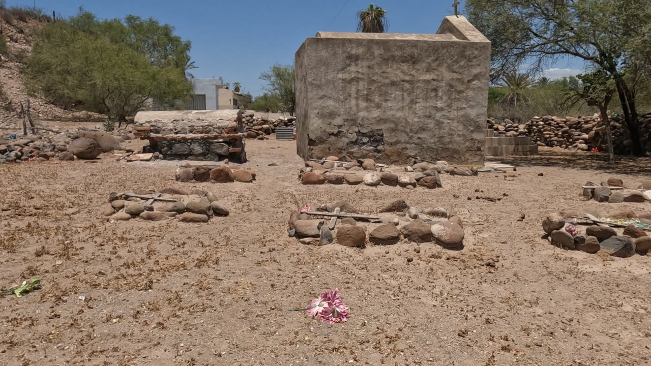 Baja California Mexico desert cemetery view with sandy ground, low stone grave outlines, sparse trees, a rough stucco wall, blue sky, and no visible people.