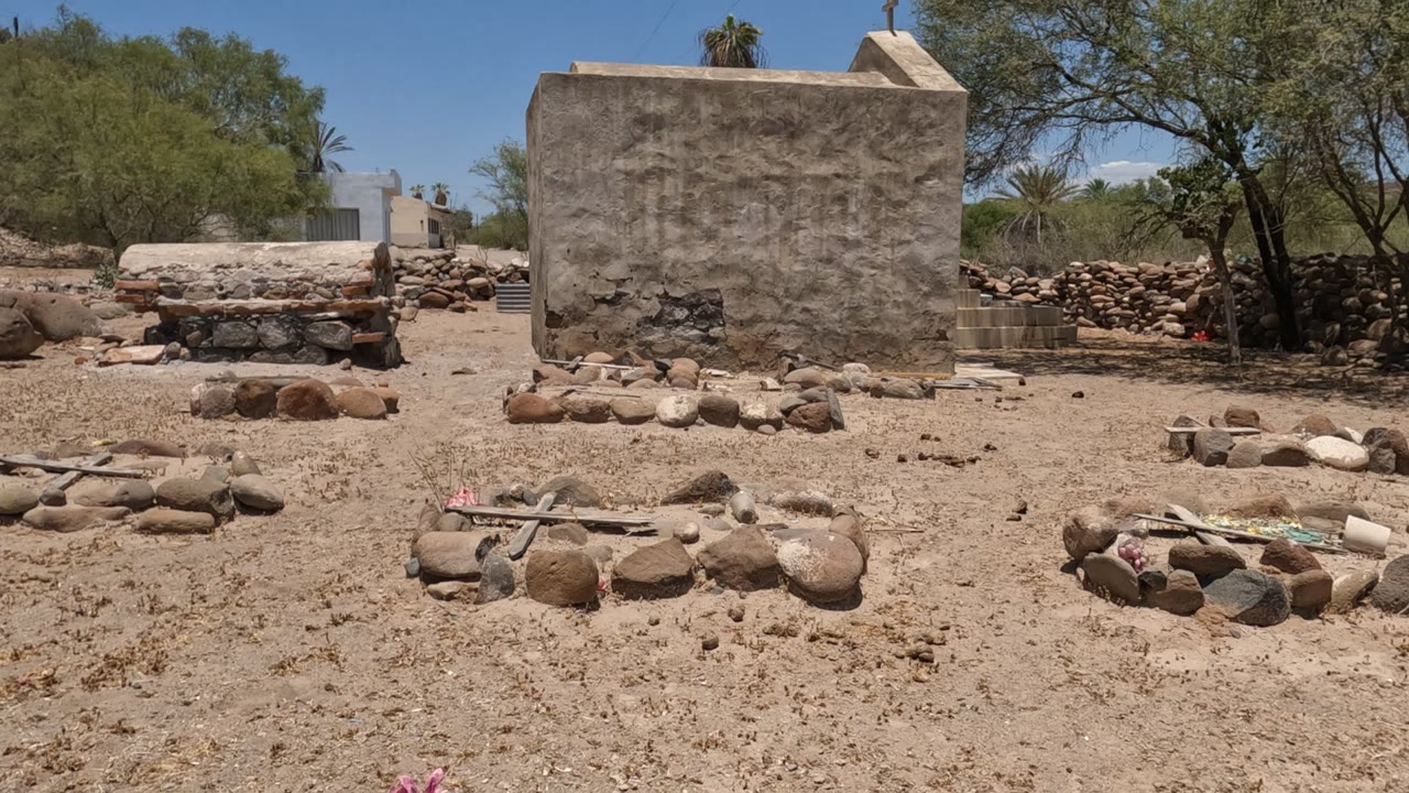 Sandy desert cemetery with low stone graves and sparse trees.