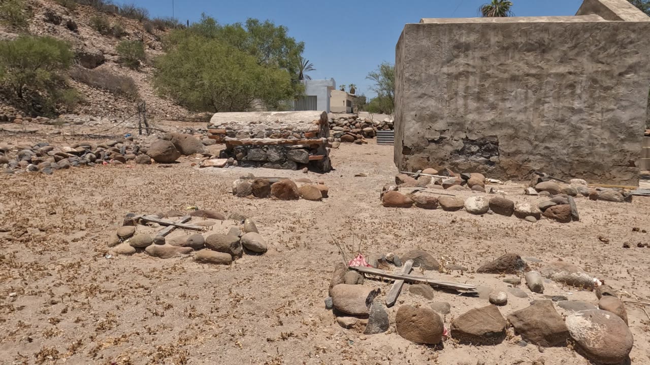 Baja California Mexico cemetery frame with a rough stucco wall centered behind stone grave outlines, scattered rocks, dry scrub, trees, and no visible people.