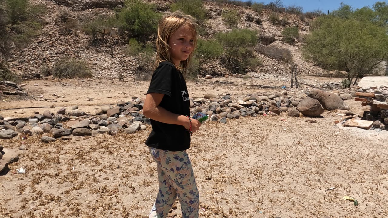 A child stands at the left edge of a dry rocky yard in Baja California Mexico, with sandy ground, scattered stones, sparse trees, and a scrubby hillside in the background.