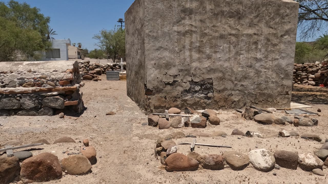 A low stone structure sits in the foreground of the Baja California Mexico yard, with a rough plaster wall to the right, scattered rocks, trees, and distant small buildings visible, no visible people.