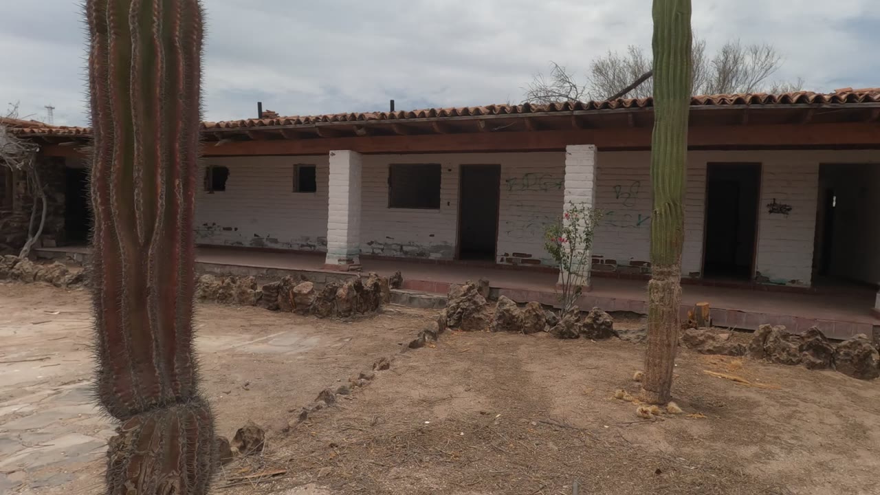 A child appears in the Baja California Mexico courtyard, walking across sandy ground near a stone border with a white adobe-style building, dark openings, red roof tiles, and tall cacti behind them.