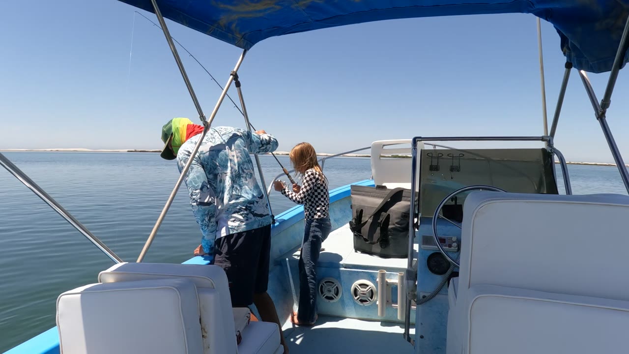 In Baja California Mexico, an adult in a blue patterned shirt and green cap leans over the rail of a blue boat while a child stands beside a fishing rod on calm water.