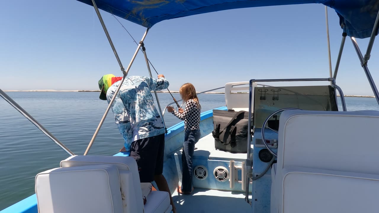 In Baja California Mexico, the adult bends toward the blue rail as the child holds or watches the fishing rod beside the console and white seats on smooth open water.