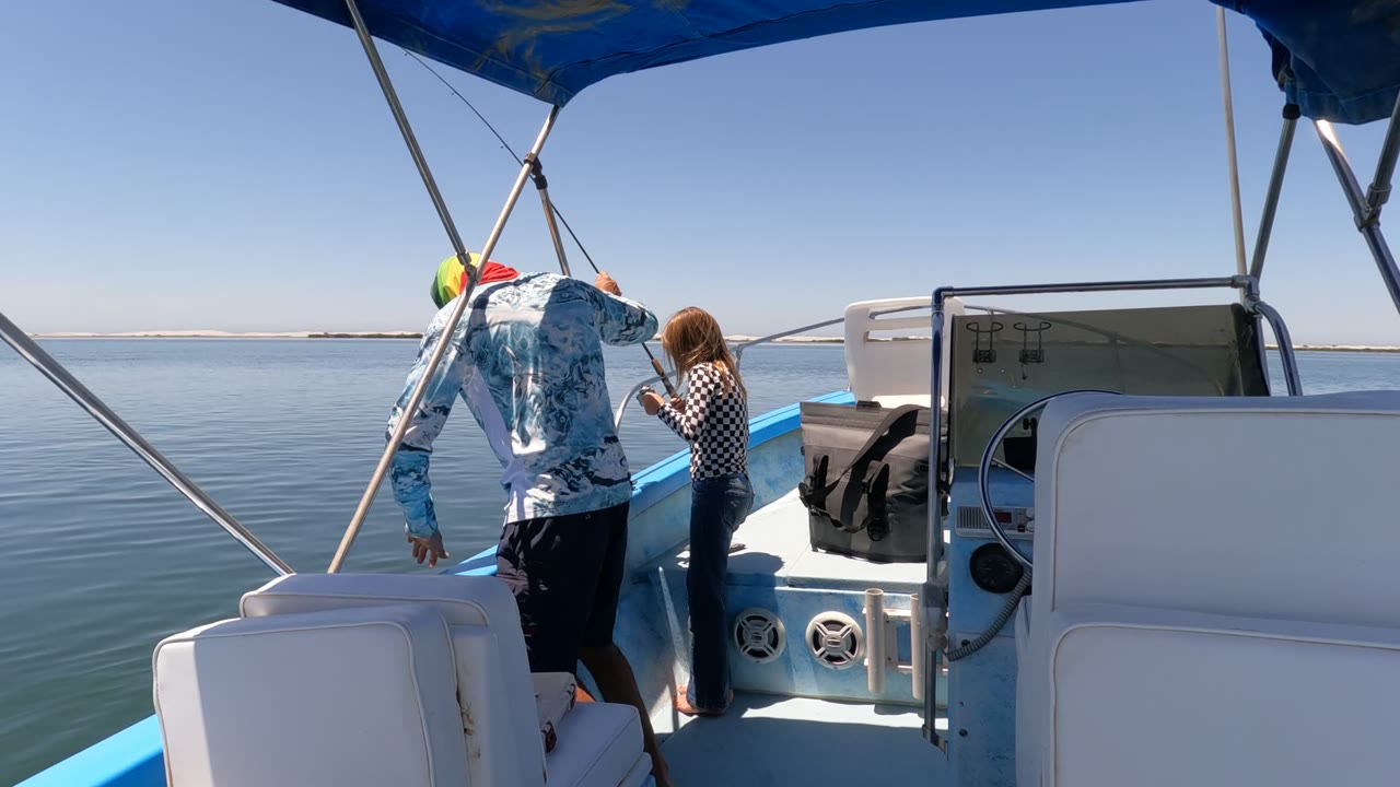 In Baja California Mexico, the adult reaches lower along the blue boat side while the child stands close to the rail with the fishing rod angled over calm water.