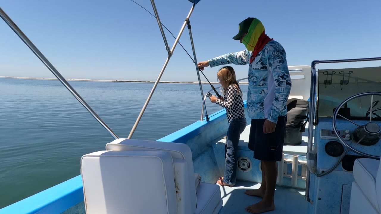 In Baja California, Mexico, an adult in a green hat stands beside a child holding a fishing rod on a blue-and-white boat over calm water.