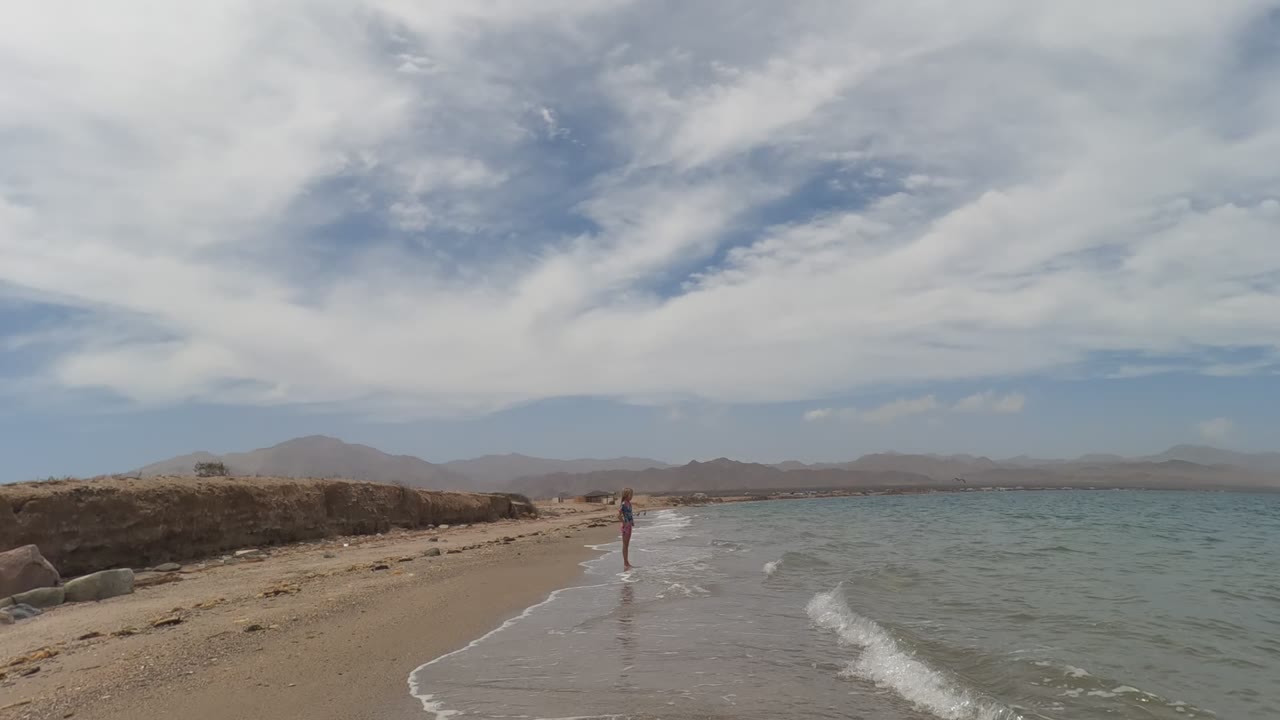 In Baja California Mexico, a person stands near the shoreline as small waves reach a sandy beach, with teal water, distant mountains, and broad white clouds overhead.