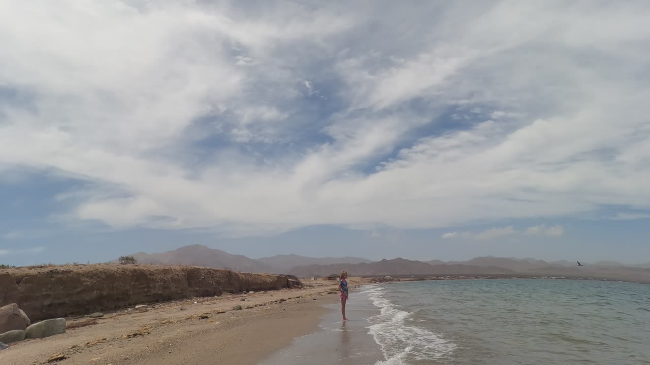 In Baja California Mexico, the person stands farther right in the wide frame beside the surf, with a sandy curve, green-blue water, low mountains, and layered white clouds visible.