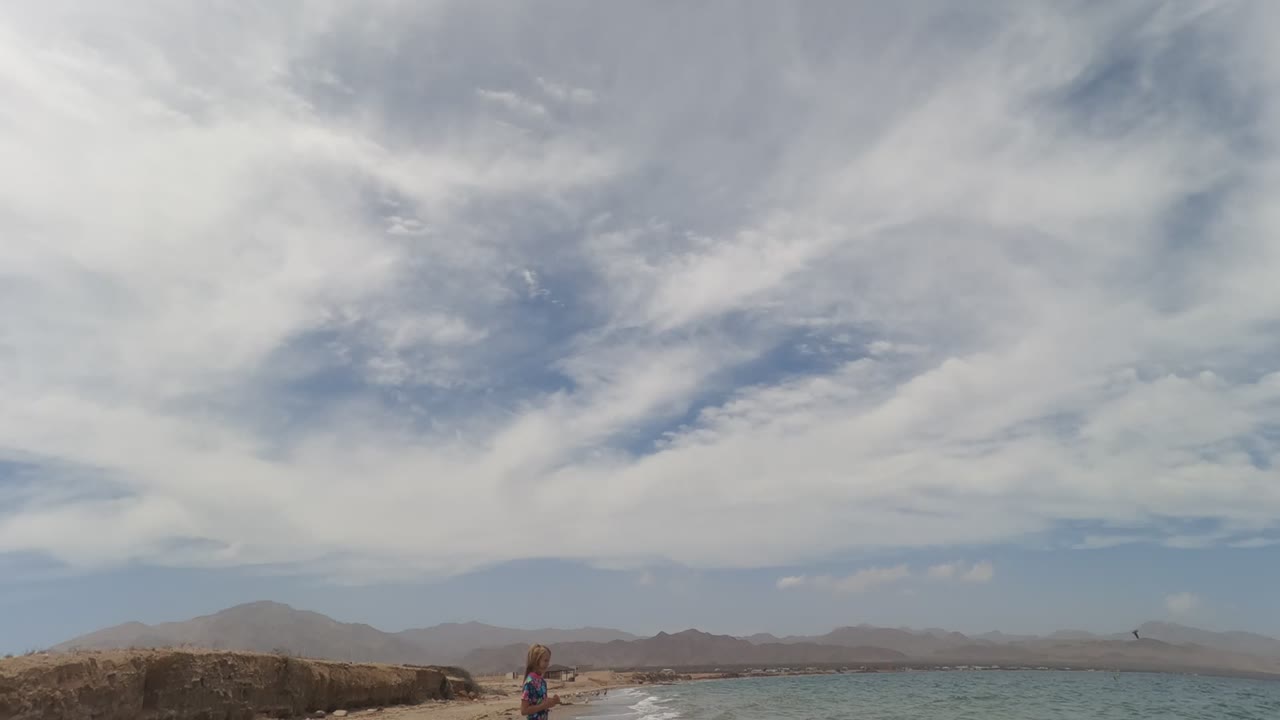 A child stands low in the frame on a sandy Baja California Mexico beach, with calm water, distant mountains, and sweeping white clouds above.