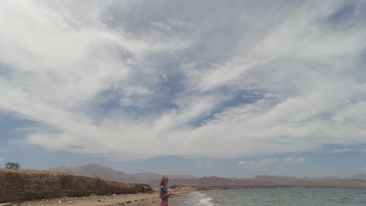 The child appears near the shoreline beside a rocky bluff, with pale blue water and hazy mountain ridges across the Baja California Mexico horizon.