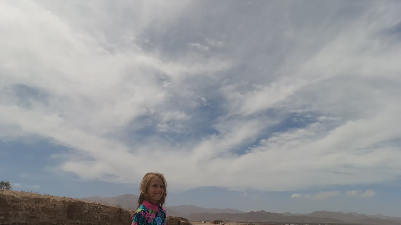 A girl in a floral shirt stands at the lower left of a Baja California Mexico coastal frame, with ocean water, low hills, and streaked clouds behind her.
