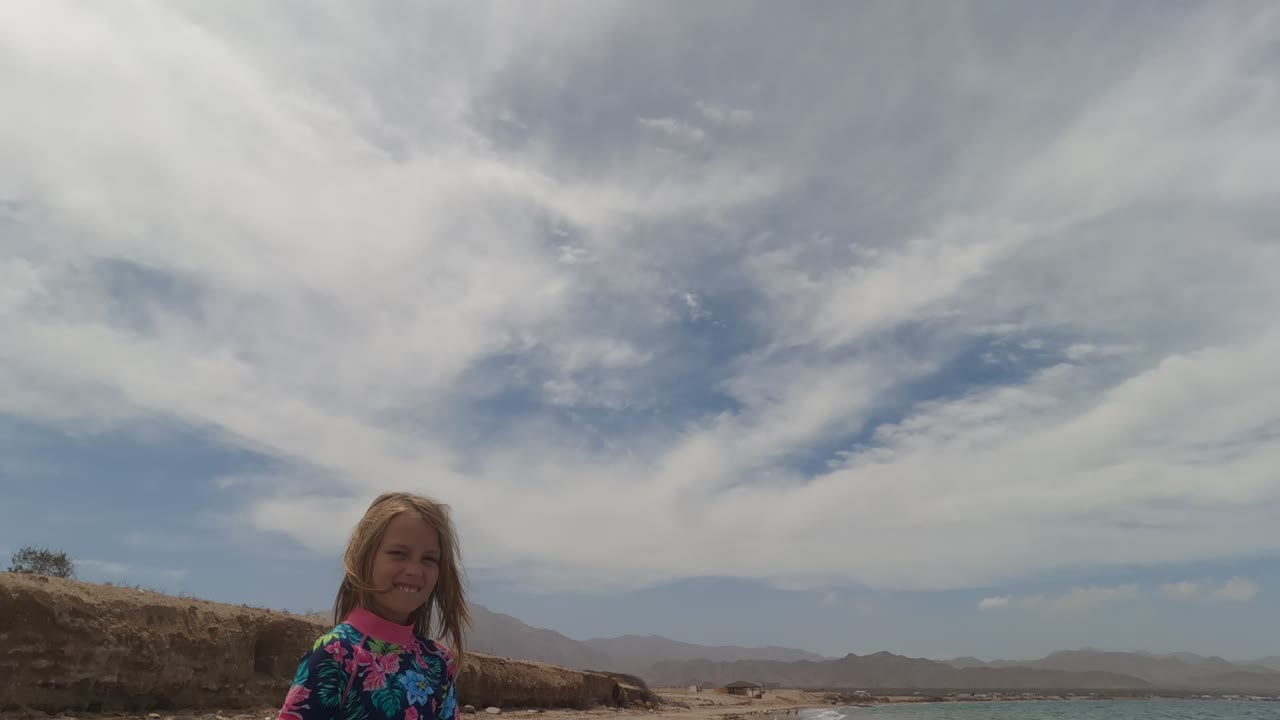 The girl appears closer and near the center of the Baja California Mexico beach scene, smiling with rocky ground, ocean, distant hills, and high white clouds visible.