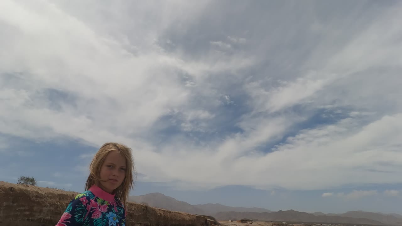 The girl stands at right of frame in Baja California Mexico, looking toward the camera with brown coastal terrain, ocean water, distant hills, and a broad cloud layer behind her.