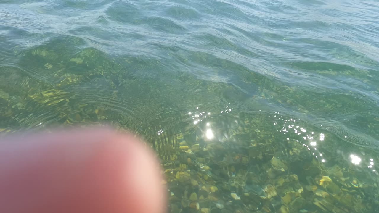Clear water in Baja California Mexico fills the frame with sunlight on ripples, seabed visible below the surface, and a fingertip obscuring the lower left foreground with no visible people.