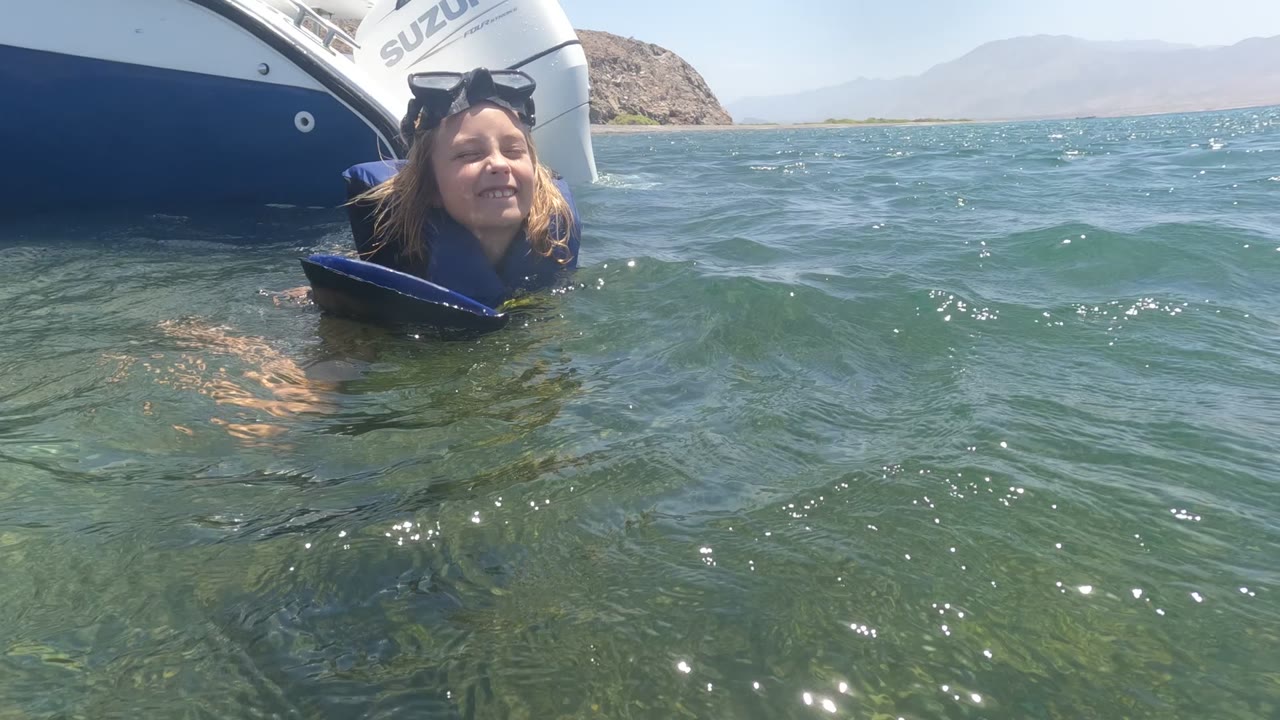 The child remains in the water in Baja California Mexico as small waves rise in front of her, with the boat hull and Suzuki motor close behind and mountains faint on the horizon.