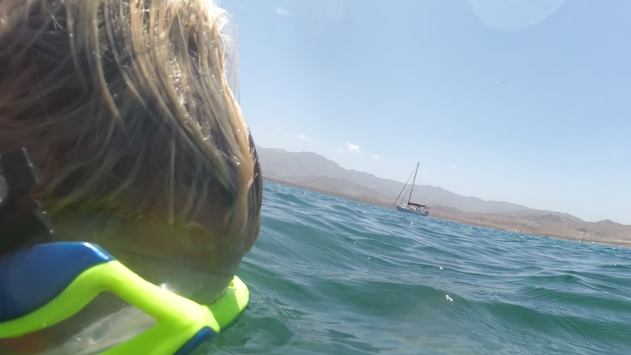 Close water-level view behind the blonde snorkeler in Baja California Mexico as she faces open water, with a sailboat, low mountains, and bright sky ahead.