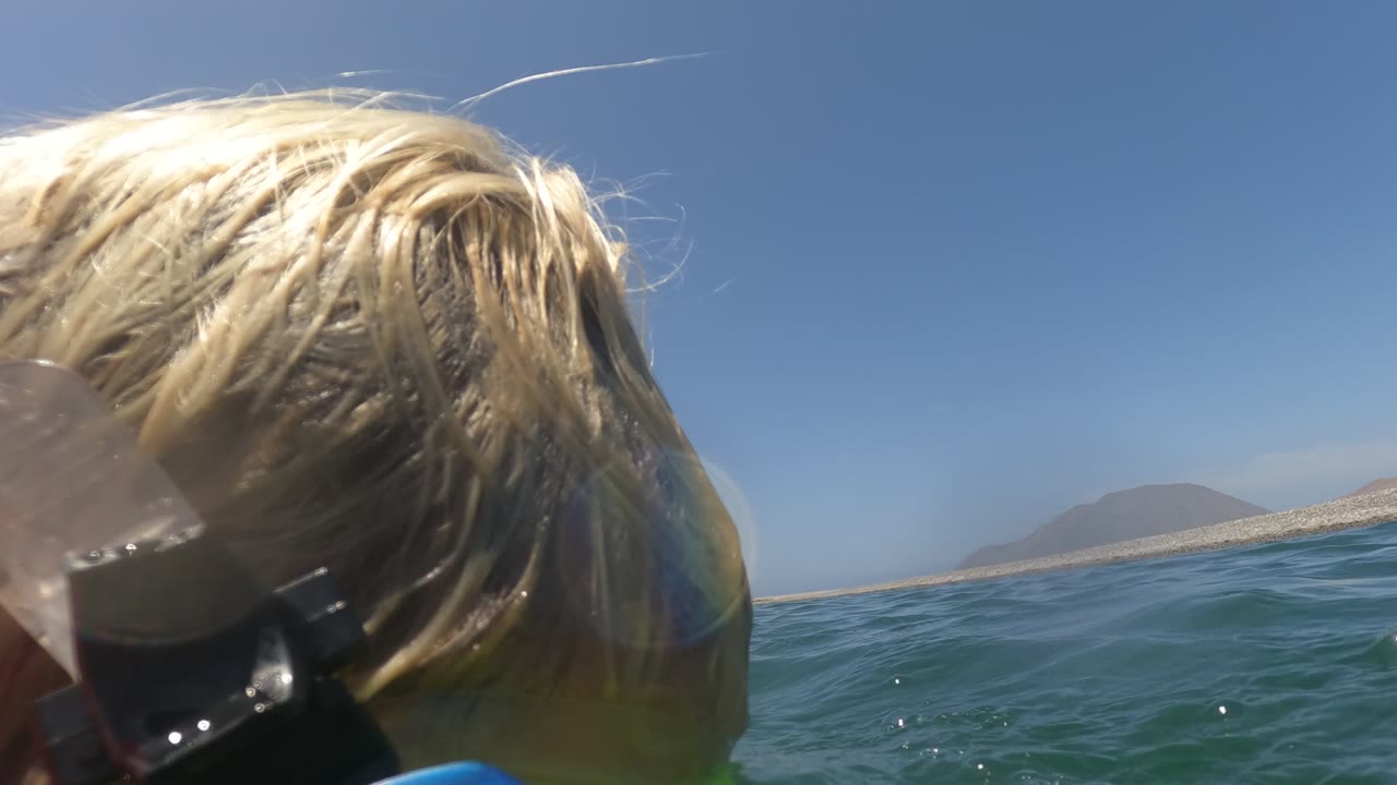Side close-up of the blonde snorkeler at the waterline in Baja California Mexico, with the snorkel mask strap visible, low waves, blue sky, and a pale mountain shoreline.