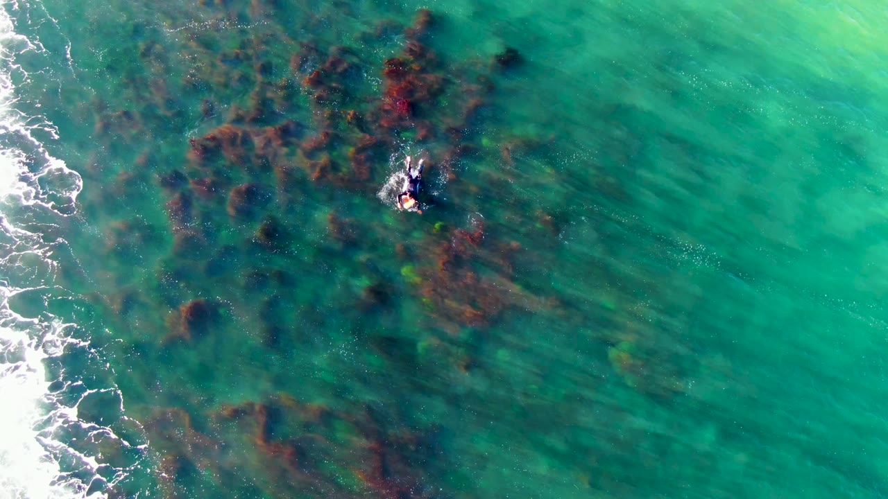 At 0:02, the La Bocana swimmer is surrounded by clearer reddish-brown underwater patches, with pale surf foam still visible along the left side.