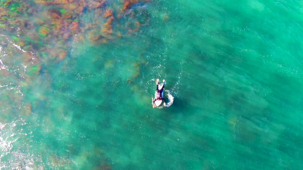 The swimmer appears farther right in the overhead La Bocana frame, surrounded by a small circular splash and broad bands of turquoise water with darker seabed marks.
