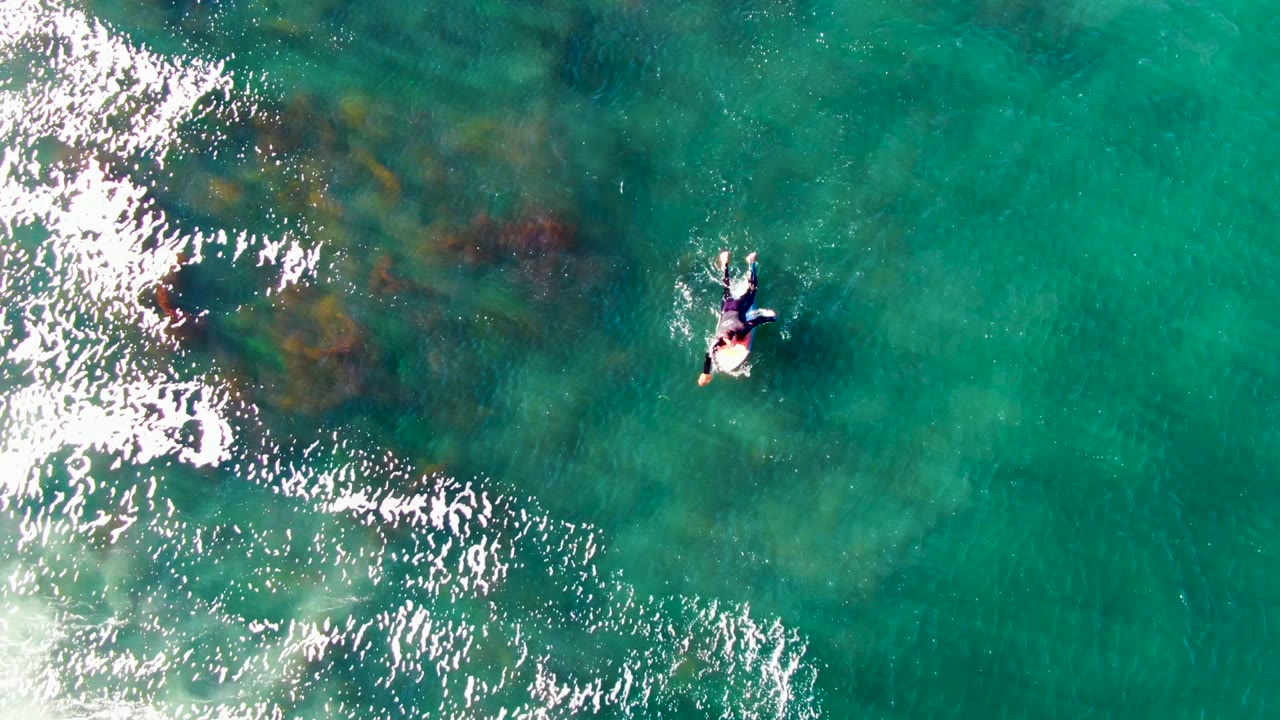 Overhead aerial frame from La Bocana, Baja California Sur showing a person on a surfboard near the upper center, with bright white foam entering from the left over clear turquoise water.