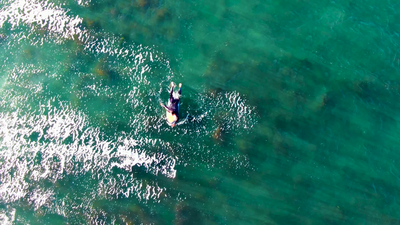 Overhead frame from La Bocana, Baja California Sur showing a person on a surfboard near bright white surface ripples in clear turquoise water.