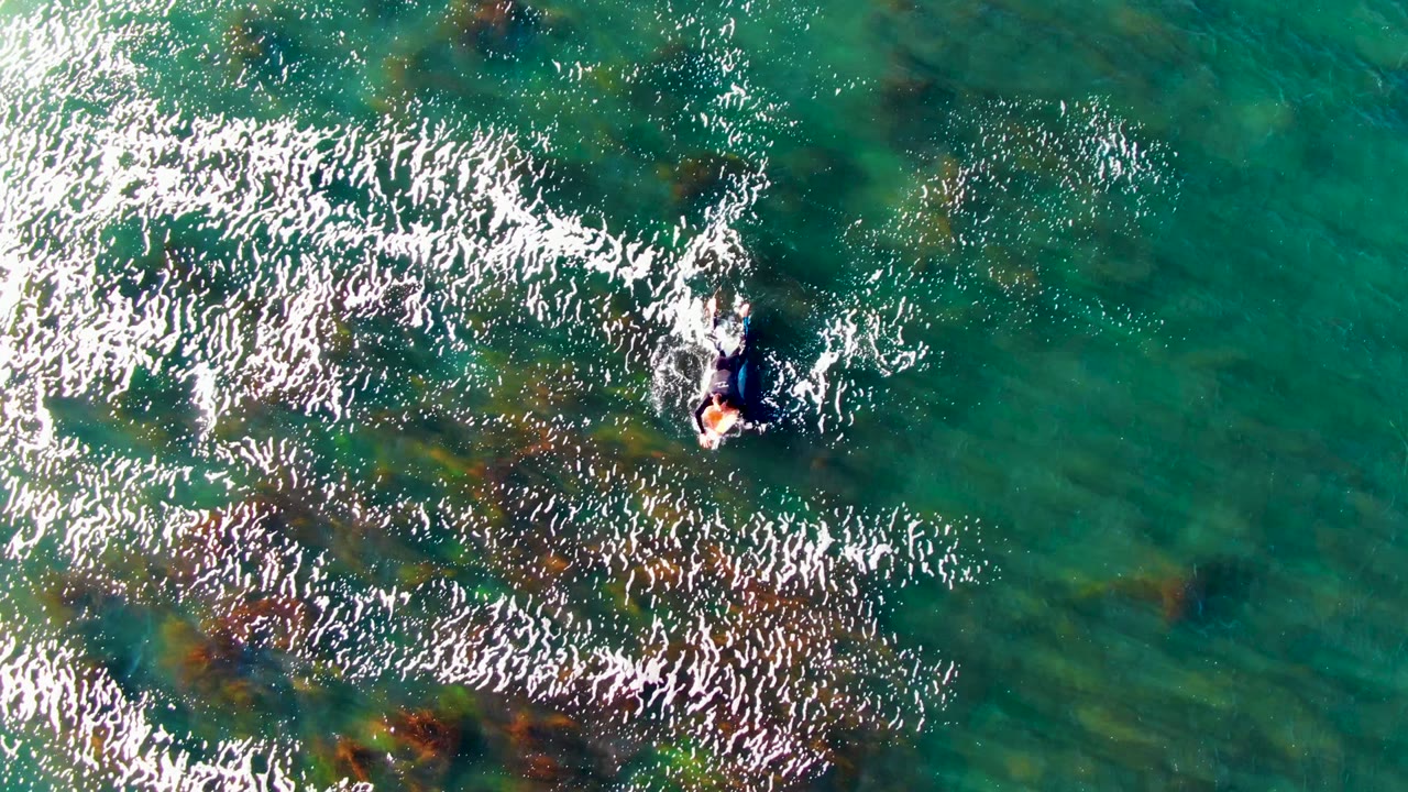 At La Bocana, Baja California Sur, Mexico, a lone swimmer appears near the upper center of clear turquoise water with white foam and dark underwater vegetation below.