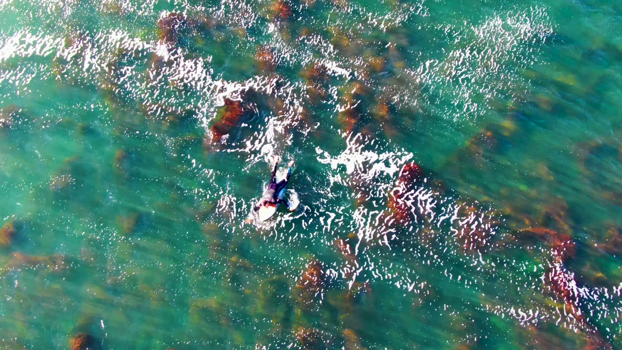 Overhead frame from La Bocana, Baja California Sur shows a swimmer near the left-center moving through shallow turquoise water with bright sparkle and dark underwater rocks.