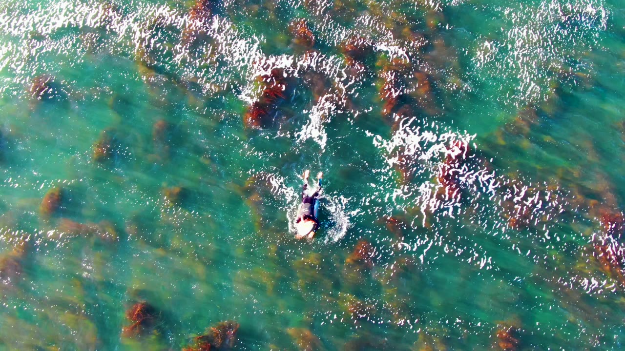 At 0:02, the swimmer appears near the center of the La Bocana frame, surrounded by clear green-blue water, foam flecks, and scattered dark rock shapes below.