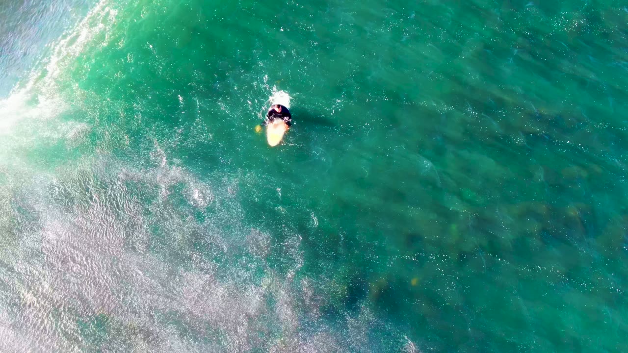 Overhead frame at La Bocana, Baja California Sur, showing a person on a pale bodyboard near a band of white surf in clear turquoise water.