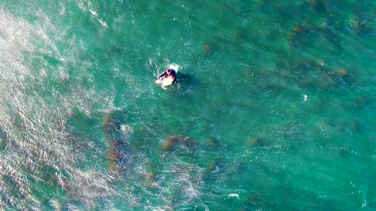 Top-down La Bocana ocean frame showing the bodyboarder small in the upper area, with whitewater sweeping in from the left and darker reef-like patches below the surface.