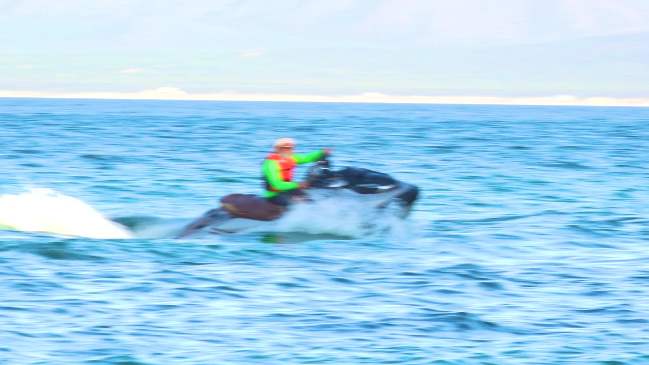 A person in a bright life jacket rides a dark personal watercraft left to right across blue ocean water, with white spray trailing behind and a pale shoreline in the distance.