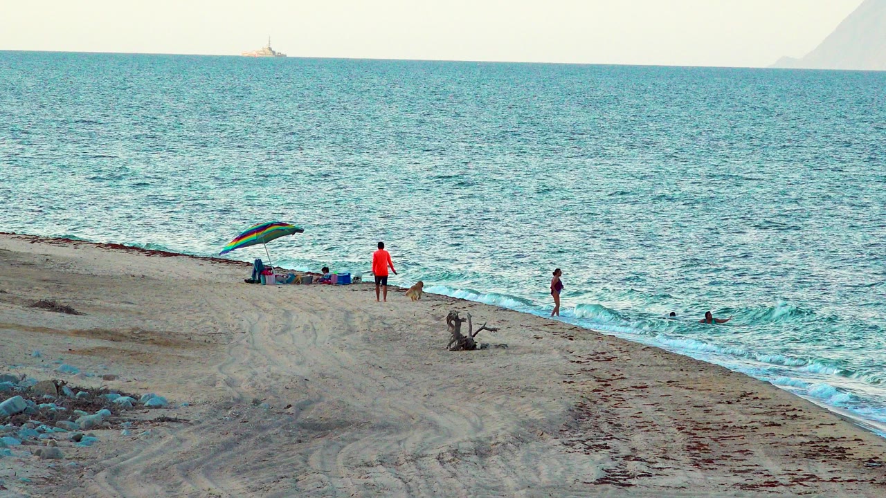 Wide view of a sandy shoreline with a striped umbrella, seated beachgoers, a person in an orange shirt, swimmers in the surf, and a small ship on the horizon.