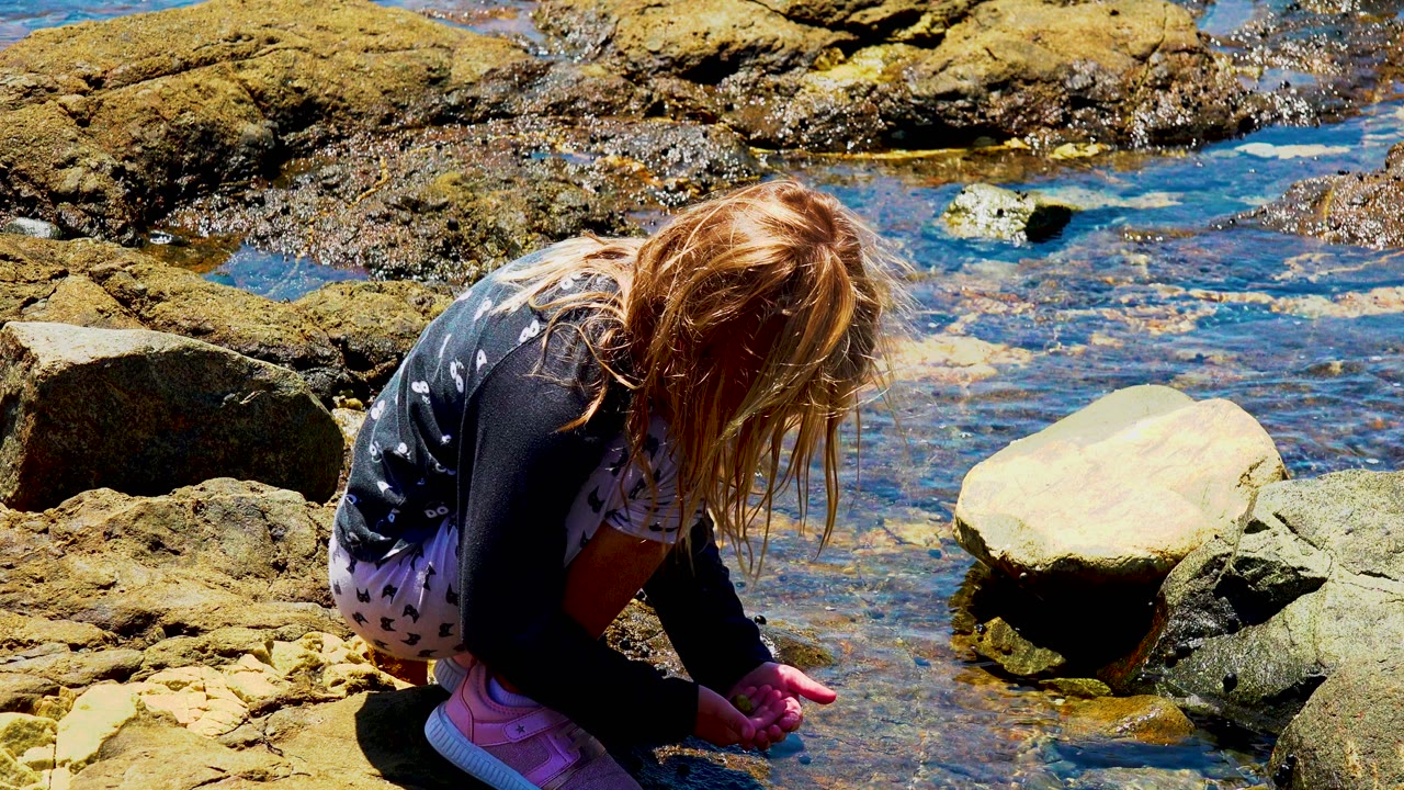 A young blonde girl crouches low beside clear shallow water and sunlit rocks at the start of the rocky shoreline clip.
