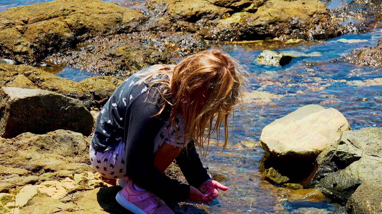 Young girl bent over rocks beside clear blue tide pool water.