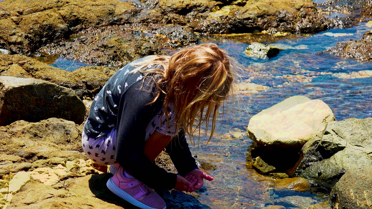 The child crouches closer to the lower left of frame while bright blue water and layered wet rocks spread behind her.
