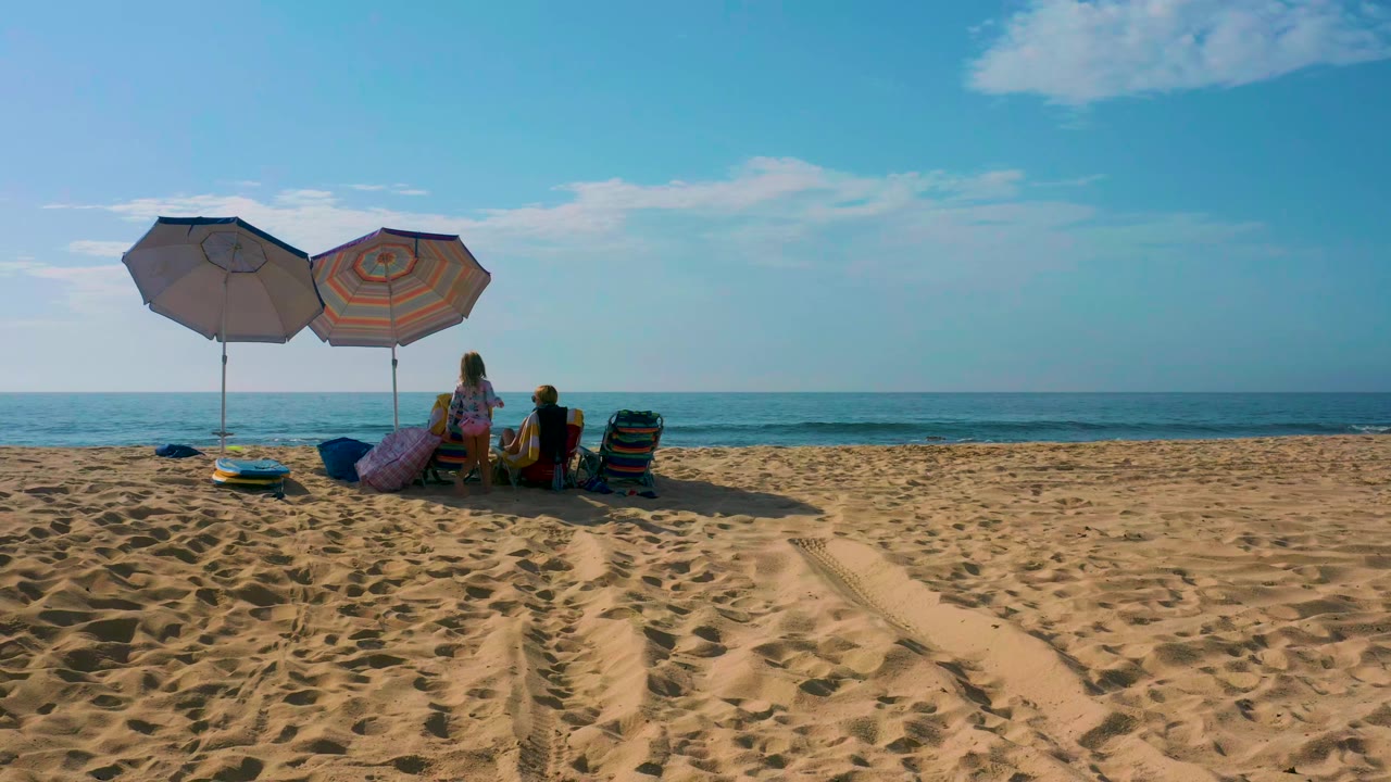 Beachgoers sit beneath two umbrellas facing the ocean.