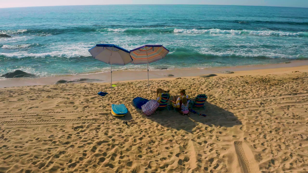 High-angle frame from Shipwrecks Beach in Baja California Sur, Mexico, showing two umbrellas shading beachgoers on textured sand with blue-green surf behind them.