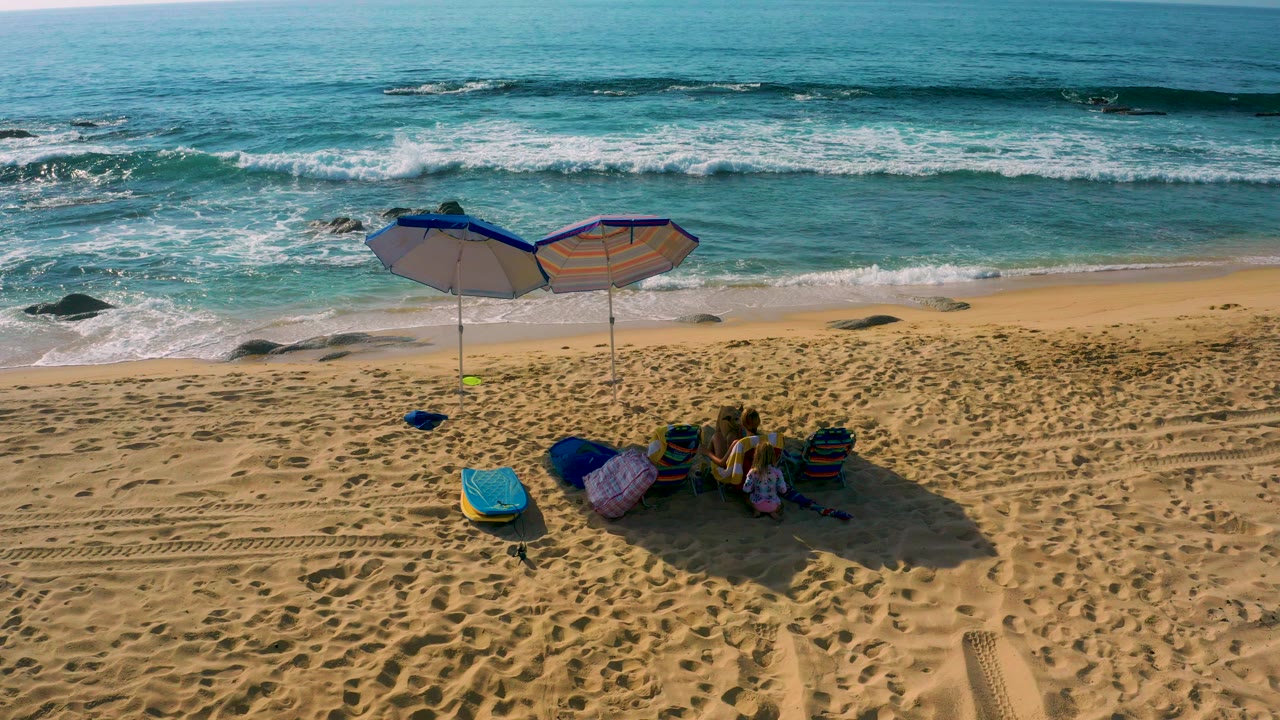 High-angle view at Shipwrecks Beach in Baja California Sur with two umbrellas, beach gear on sand, small beachgoers under shade, and white surf beyond.