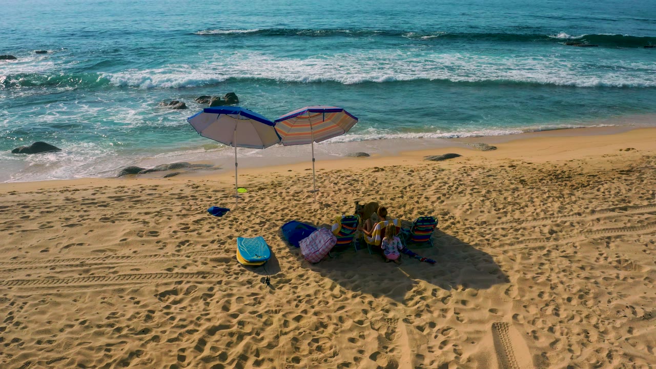Drone view over Shipwrecks Beach showing the umbrellas slightly left of center, beach items grouped in their shadow, and surf spreading across the upper frame.